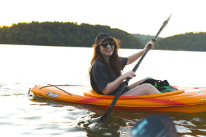 Woman wearing a baseball hat and Nöz sunscreen in an orange canoe out on a lake surrounded by hills.